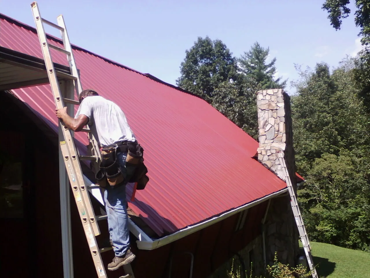 Roofer on a ladder beside a red metal roof and stone chimney — Soffit & Fascia Repair by Roofing Brothers in Vermilion